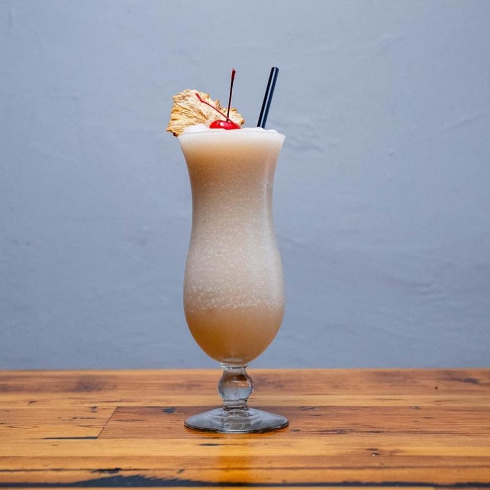 A frozen cocktail in a hurricane glass, garnished with a cherry, dried pineapple, and two straws.