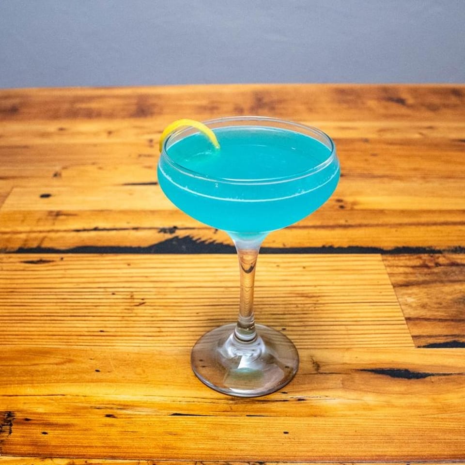 Vibrant blue cocktail in a coupe glass, garnished with a lemon twist on a wooden table.