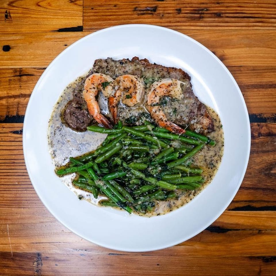 Steak and shrimp with green beans in a creamy sauce on a white plate atop a wooden table.