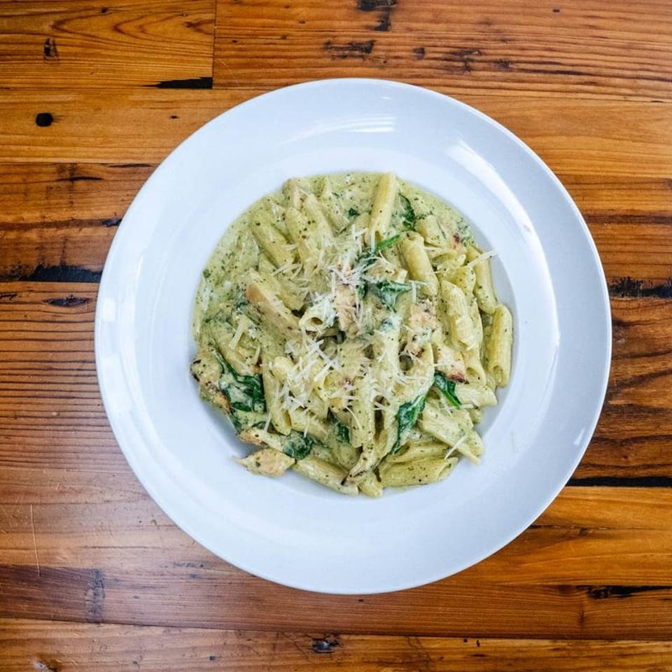 Penne pasta with creamy pesto sauce, chicken, spinach, and grated Parmesan cheese on a white plate.