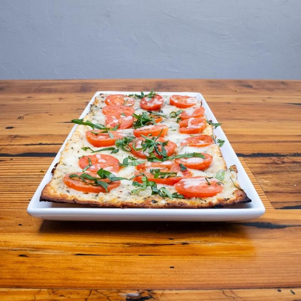 Rectangular flatbread pizza with melted cheese, sliced tomatoes, and fresh basil on a wooden table.
